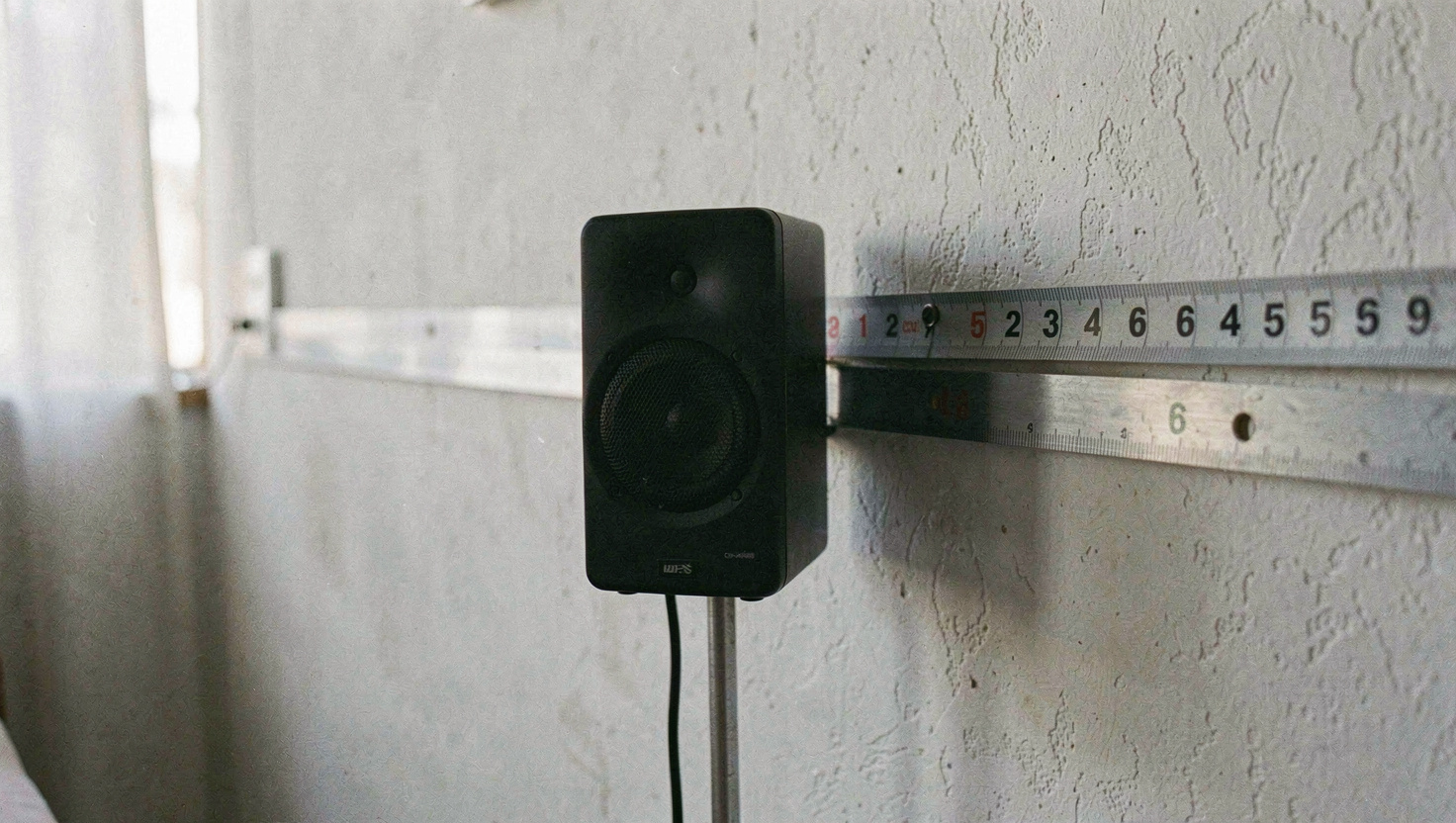 Kompaktlautsprecher auf einem schmalen Metallständer, dahinter die Wand mit angelegtem Massband, das den Abstand zeigt, kühles Tageslicht von der Seite, halbnahe Aufnahme