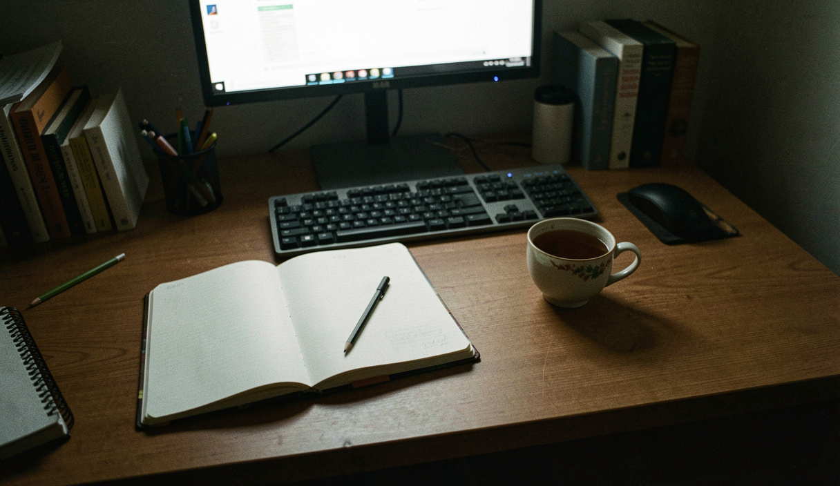 Aufgeräumter Schreibtisch mit offenem Notizbuch, Bleistift und halbvoller Teetasse, Bildschirmlicht von vorn, Zimmer sonst dunkel, Aufsicht aus Sitzhöhe