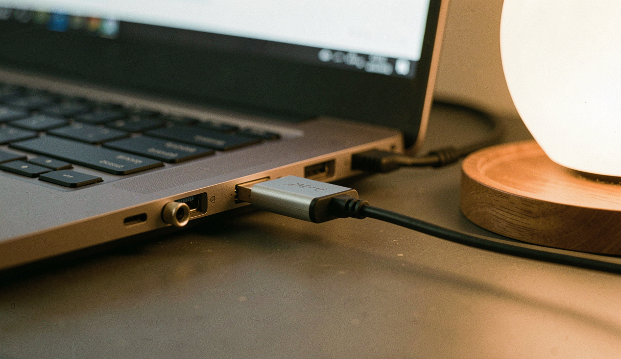 Kleiner USB-Adapter mit kurzem Kabel hängt am Anschluss eines Notebooks, daneben ein aufgeschraubter Kopfhöreranschluss, Bildschirmlicht und eine warme Lampe von rechts, Nahaufnahme